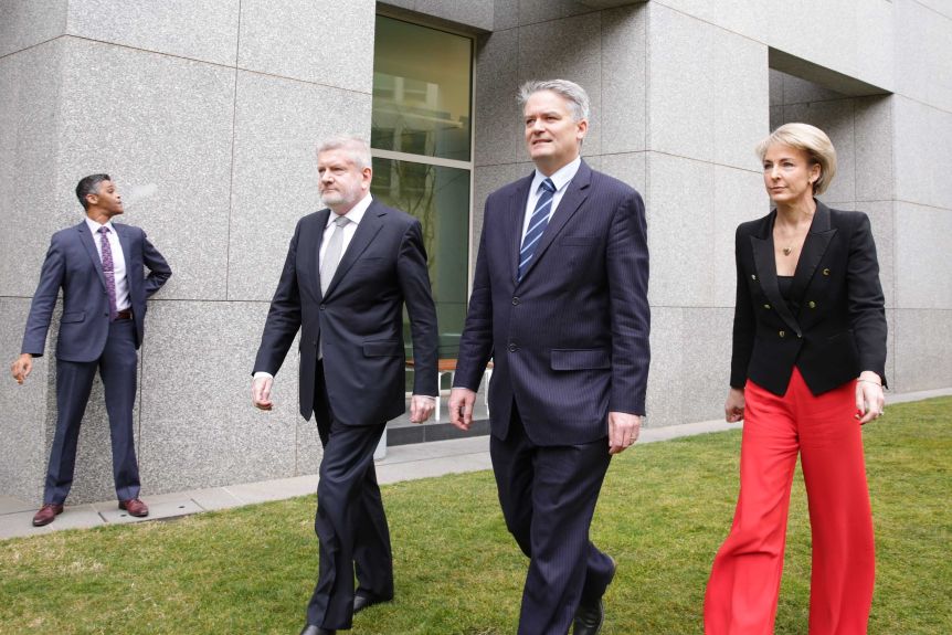 Staring straight ahead, past the camera, three Liberal powerbrokers stride past an unknown man in the background smoking.